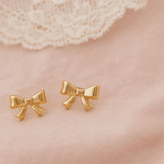 Gold bow-shaped earrings on a soft pink fabric background close up