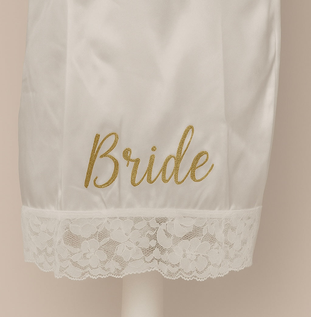 White satin robe with gold 'Bride' embroidery and lace trim on a beige background